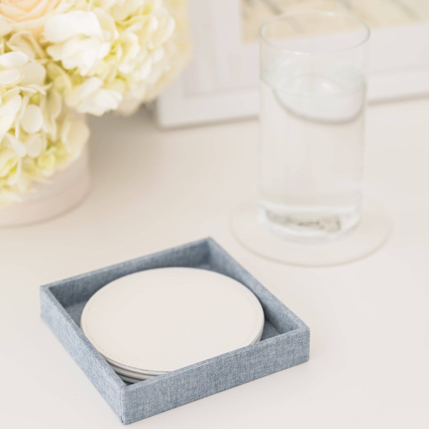 Set of white coasters in a chambray box on a light surface with flowers in the background