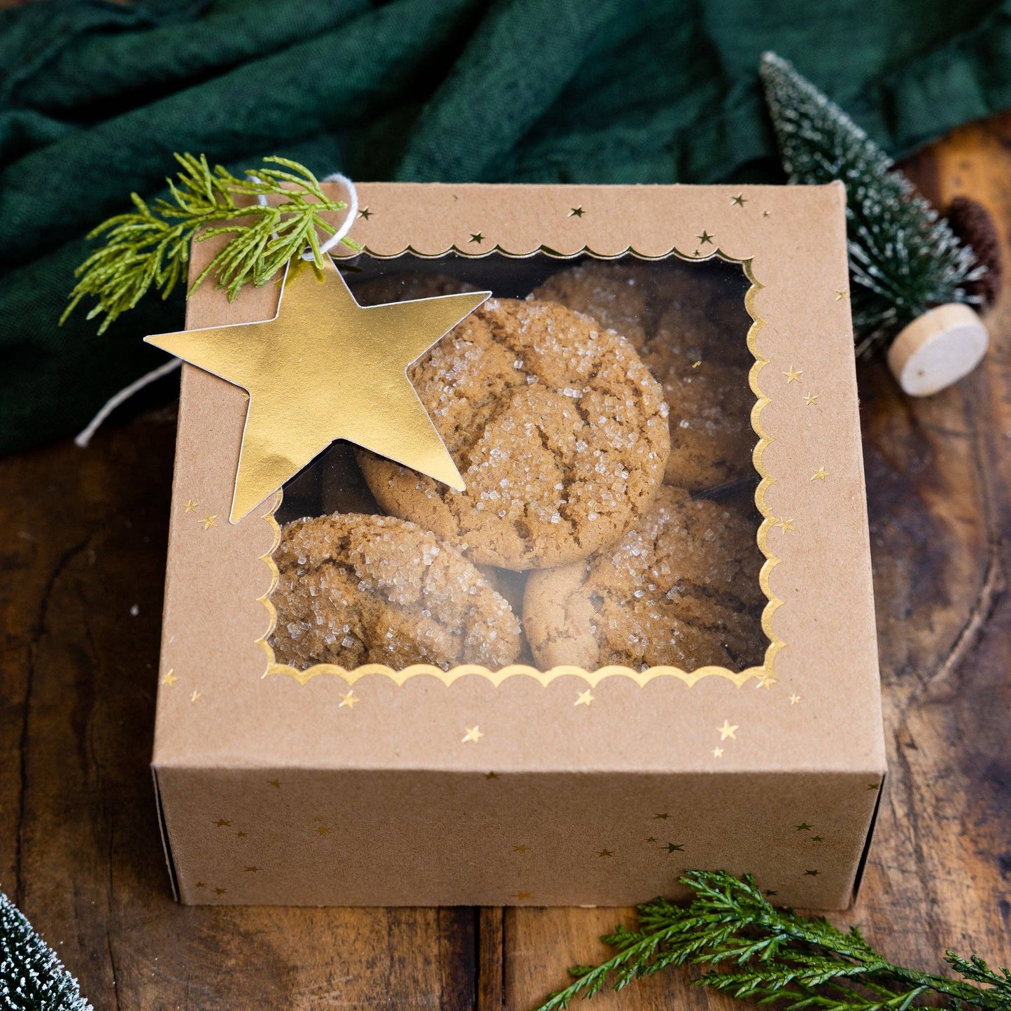 Box of cookies with a gold star gift tag on a wooden table with greenery.