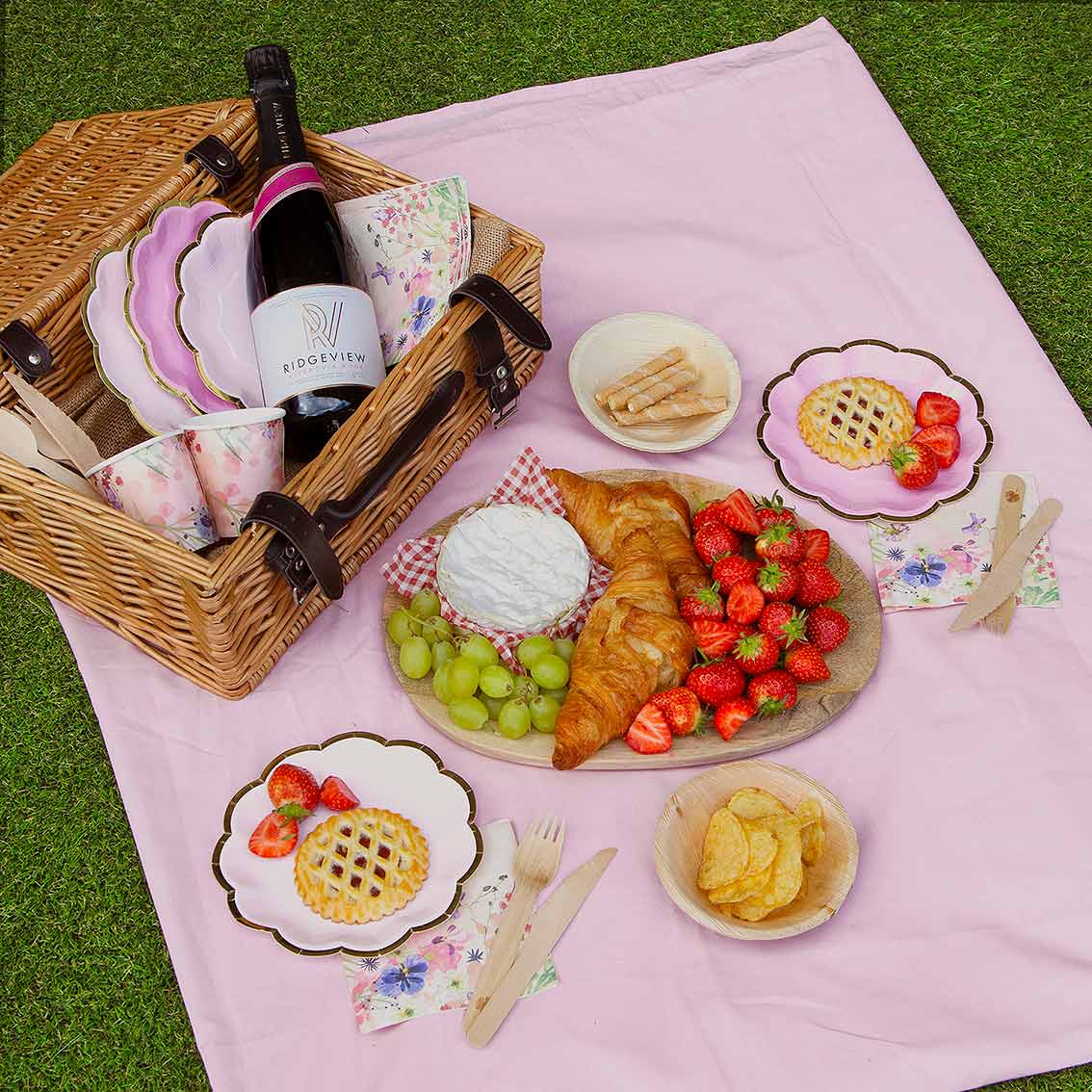 Picnic setup with a pink blanket, wicker basket, food and drinks on grass.