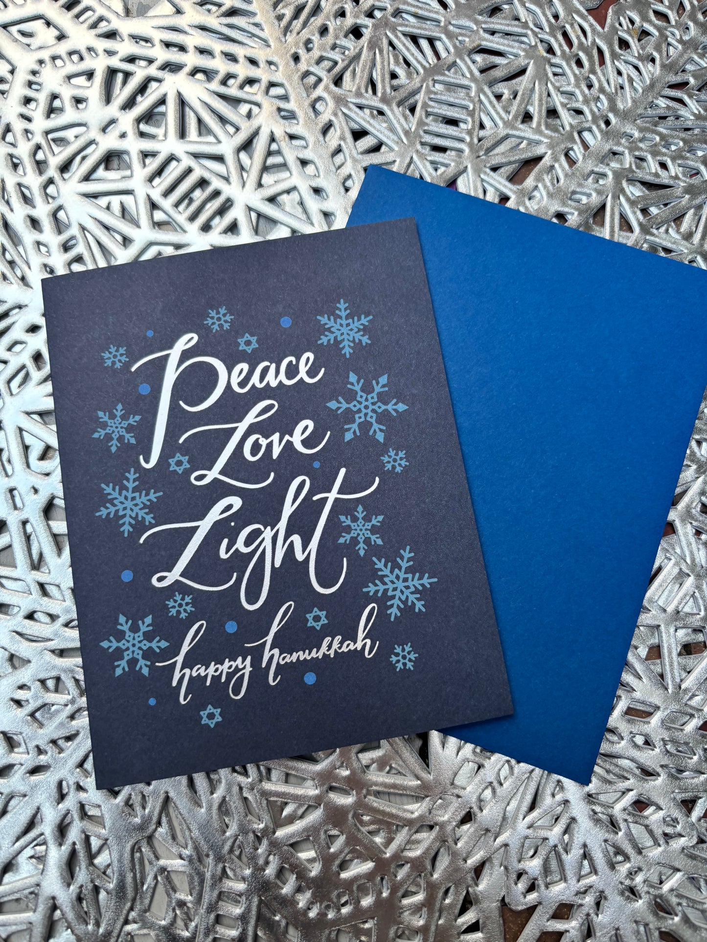 Image of Paper Source Hanukkah Card that reads "Peace, love, light, Happy Hanukkah" on a silver snowflake background