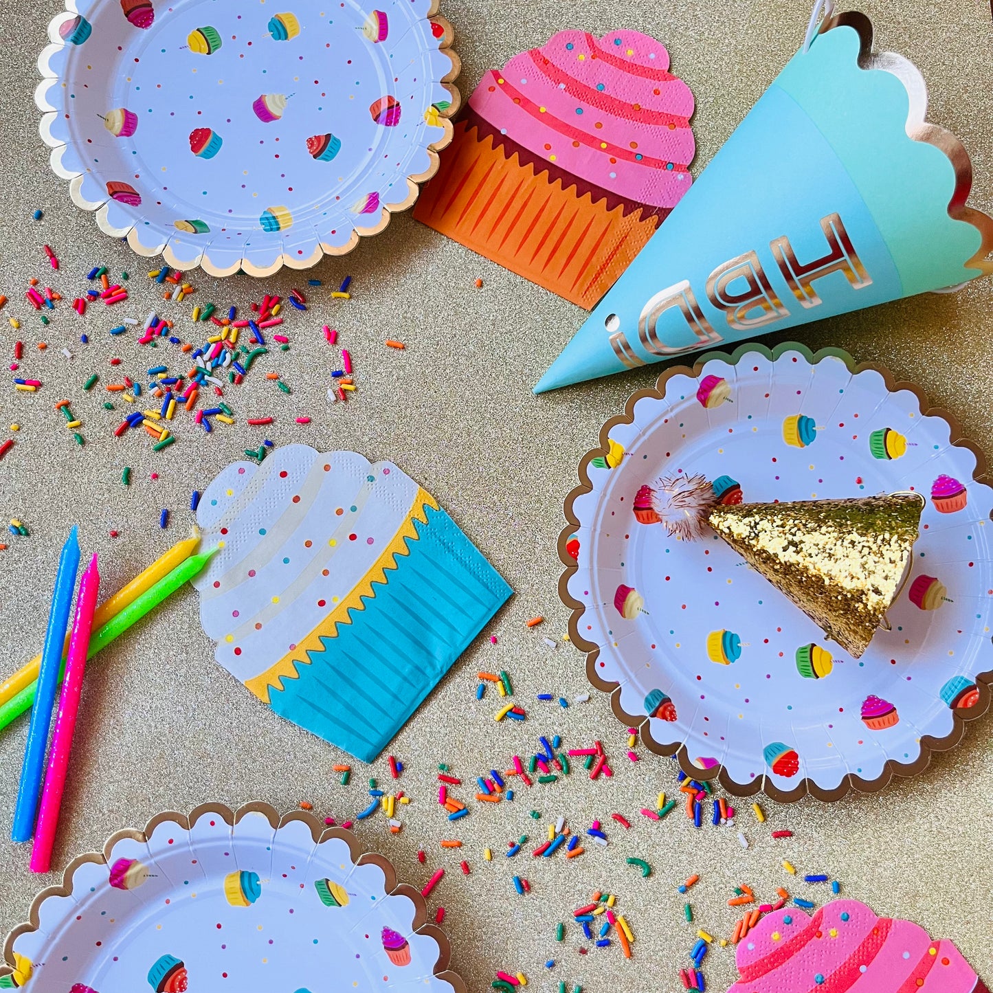 Colorful party supplies including paper plates, cups, and a 'Happy Birthday' hat on a gold glitter background with sprinkles.