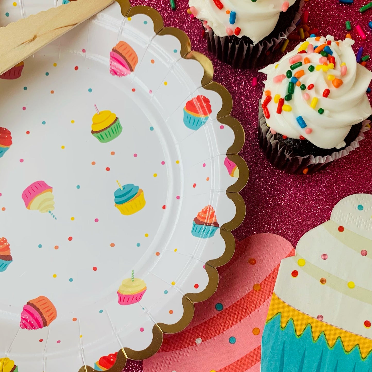 Colorful cupcake-themed paper plates and napkins on a pink glitter background with sprinkles.
