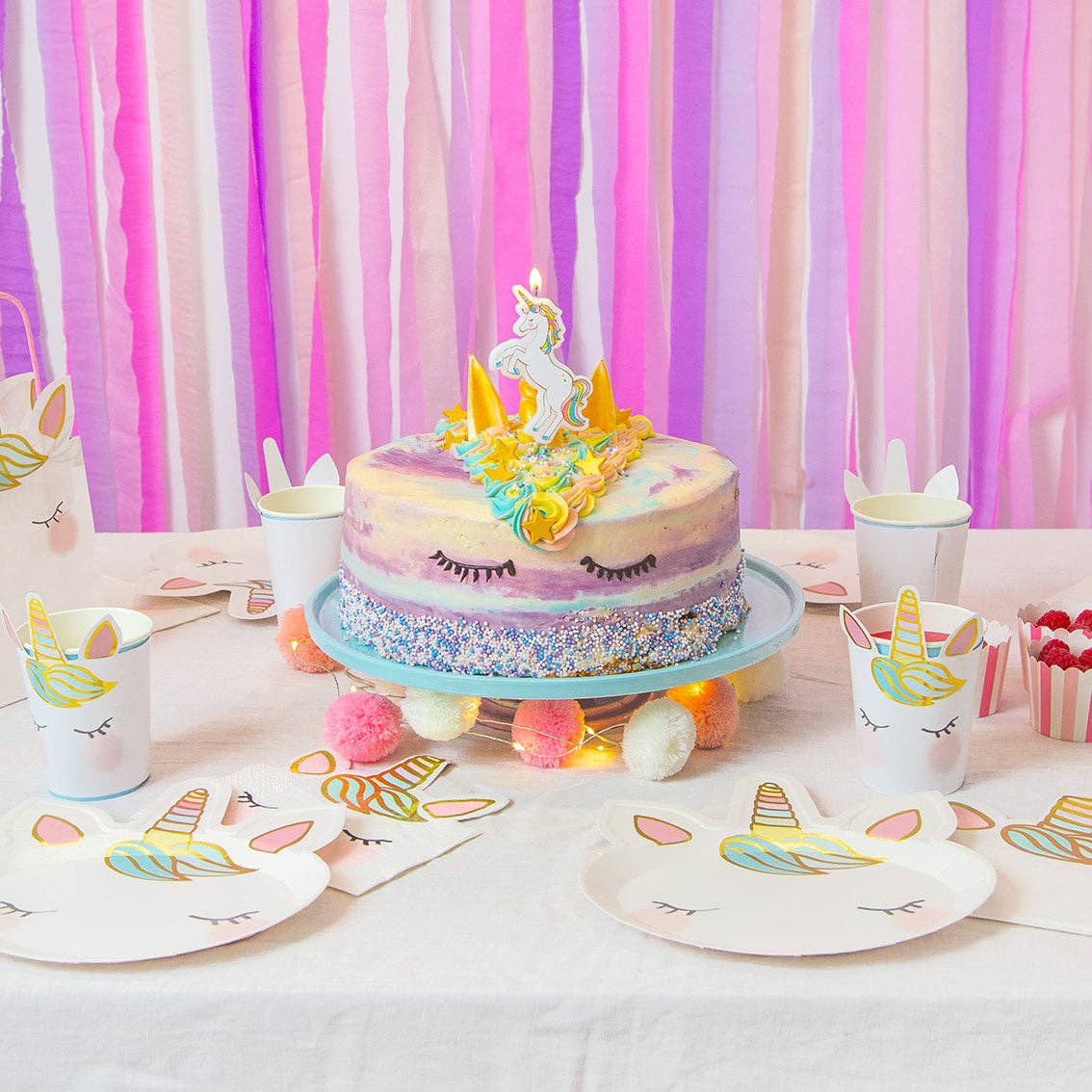 Unicorn-themed birthday party setup with a cake, plates,and cups against a colorful striped backdrop.