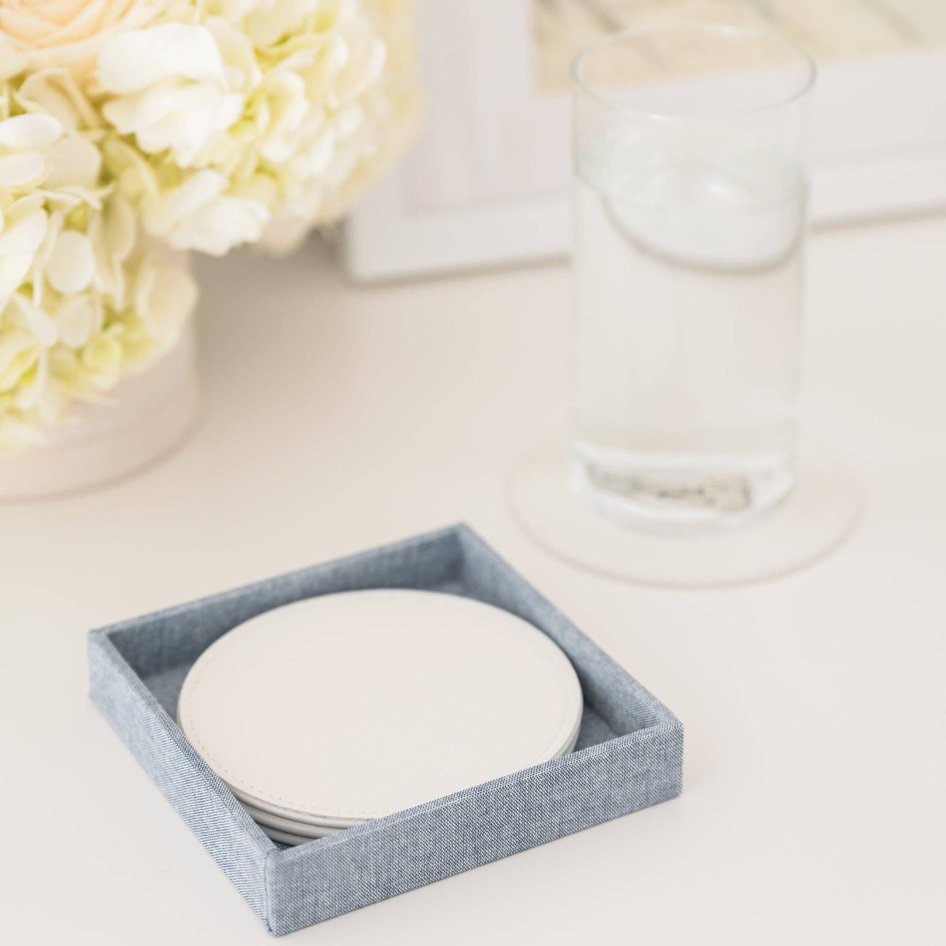 Set of white coasters in a chambray box on a light surface with flowers in the background