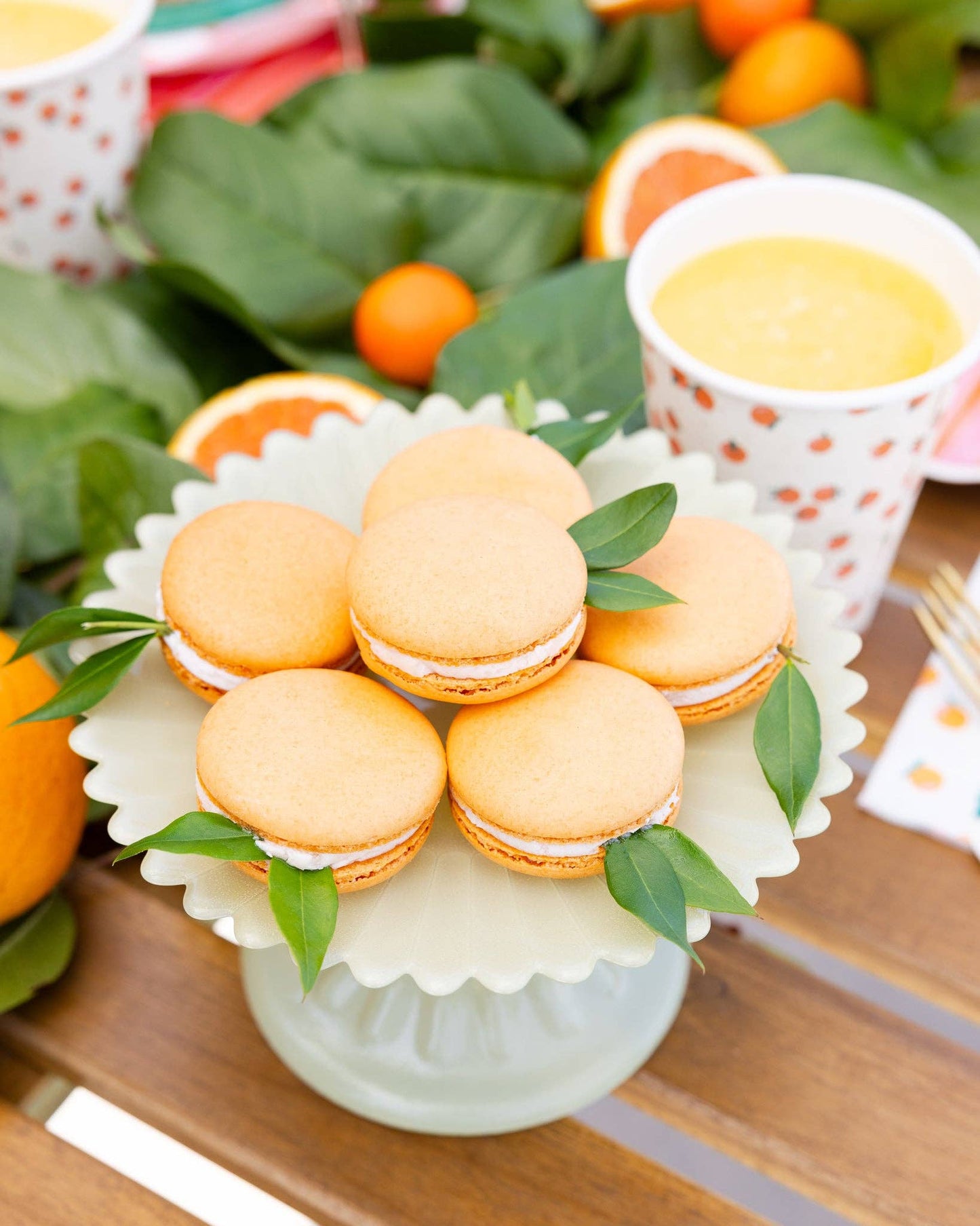 Orange macarons on a dessert stand with green leaves, surrounded by oranges and themed cups.