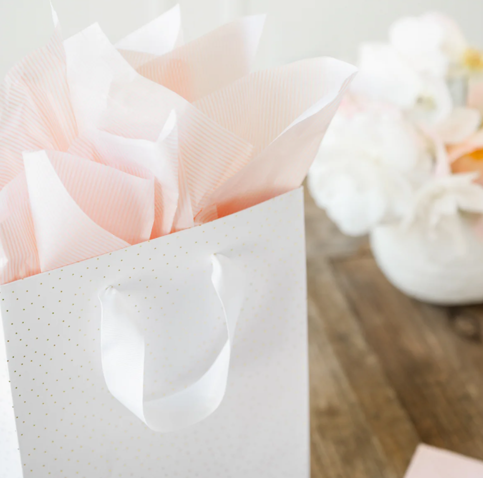 White gift bag with gold polka dots with pink tissue paper and flowers on a wooden surface