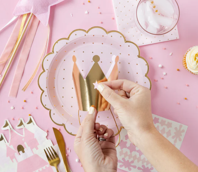 Pink and gold princess party supplies including plates, cutlery and a hand holding a candle.