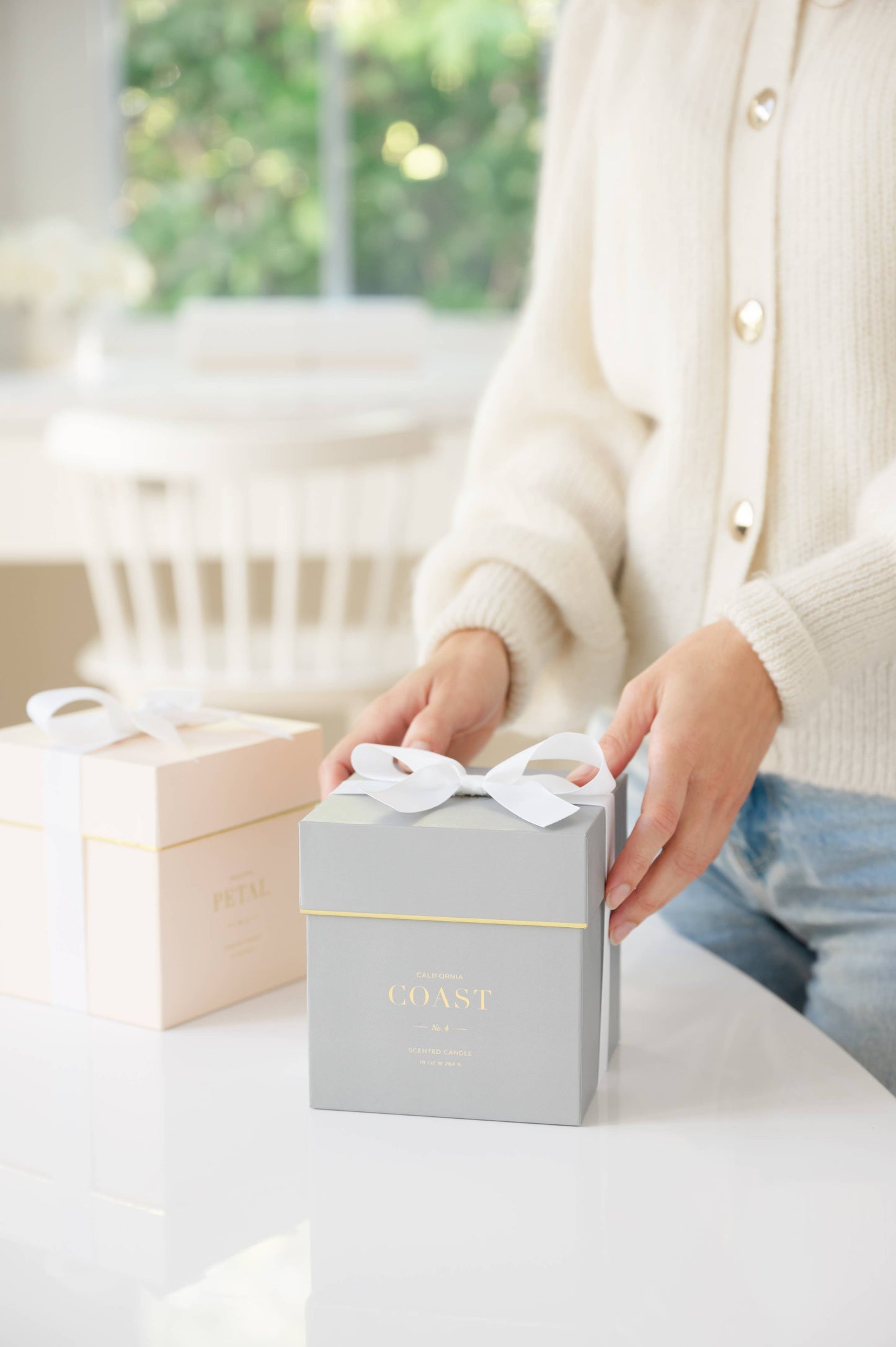 Person holding a gray candle gift box with a white ribbon in a bright room.