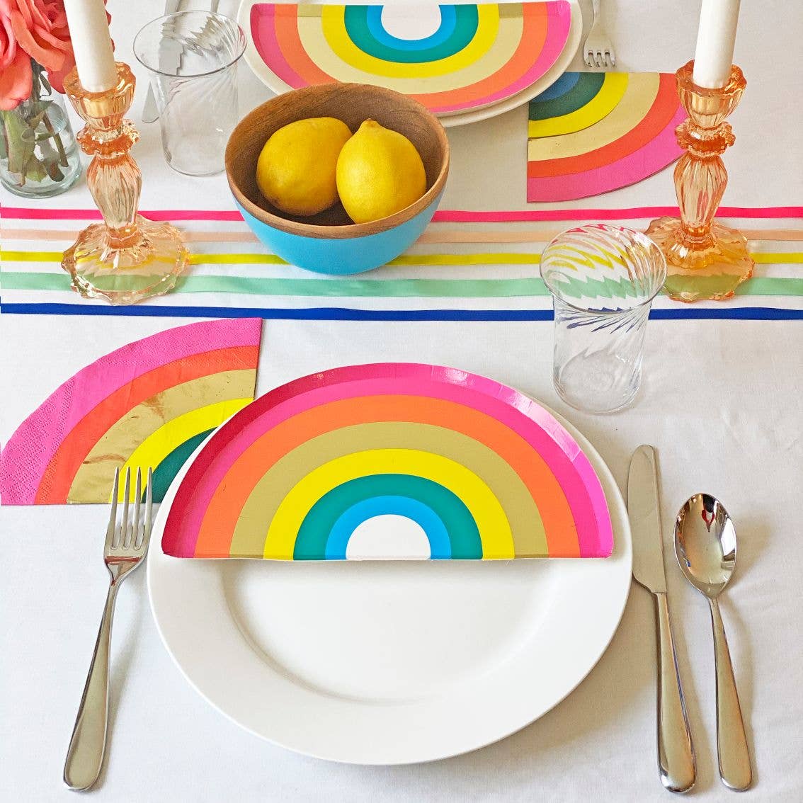 Colorful rainbow-themed table setting with plates, napkins and cutlery on a white tablecloth.